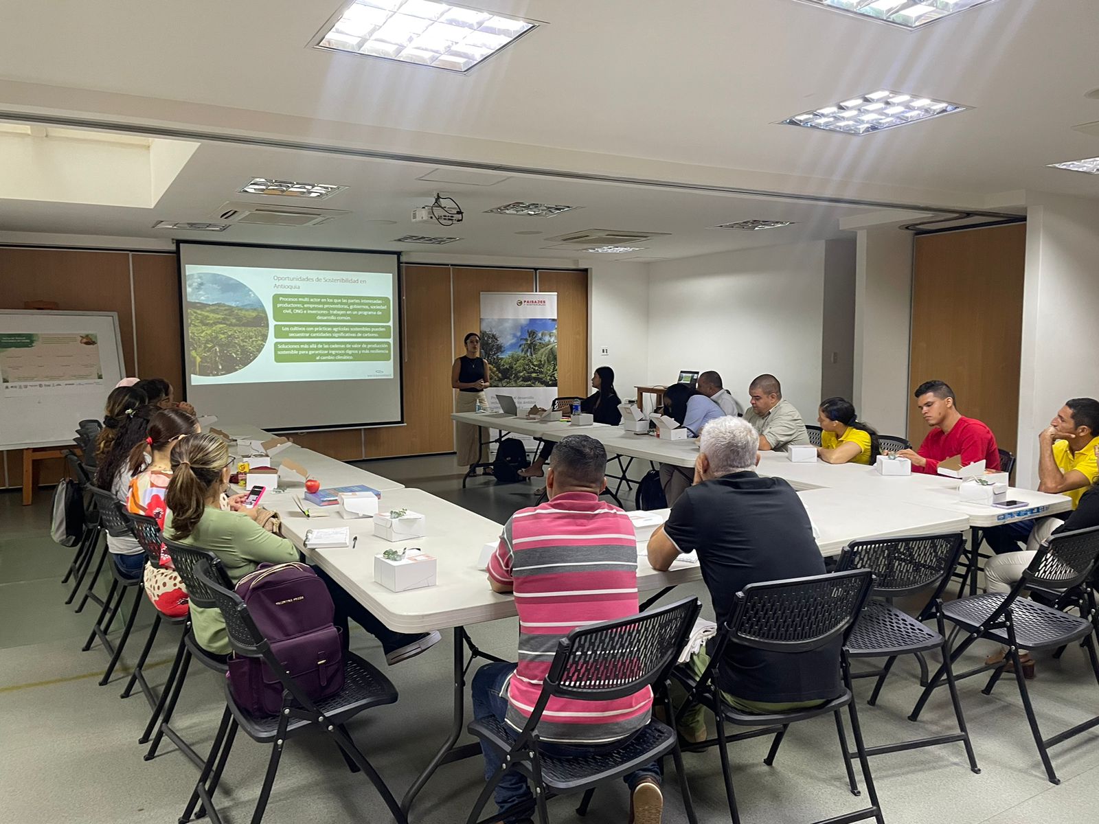 Approx. 15 people sit around white tables, watching a presentation